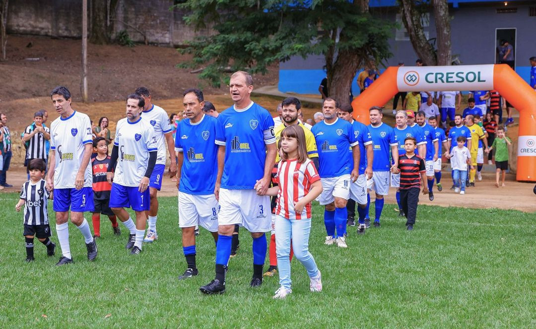 Fotografia horizontal em plano médio mostrando a entrada dos jogadores no gramado do estádio. Alinhados em fila, ex-atletas vestindo uniformes brancos e azuis caminham em direção ao campo. À frente, um homem de uniforme azul segura a mão de uma menina que veste uma camisa listrada em vermelho e branco. Ao fundo, vê-se um portal inflável na cor laranja com a inscrição 