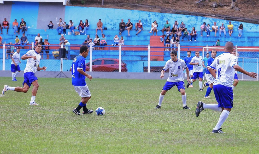 Fotografia horizontal de ação durante a partida de futebol. No centro do gramado verde, Mazinho, com uniforme azul, domina a bola com o pé direito. Ele é cercado por jogadores com uniformes brancos em posição de marcação, em destaque Zé Afonso e Mauro Galvão. Ao fundo, a arquibancada do estádio pintada em azul exibe torcedores sentados acompanhando o jogo.