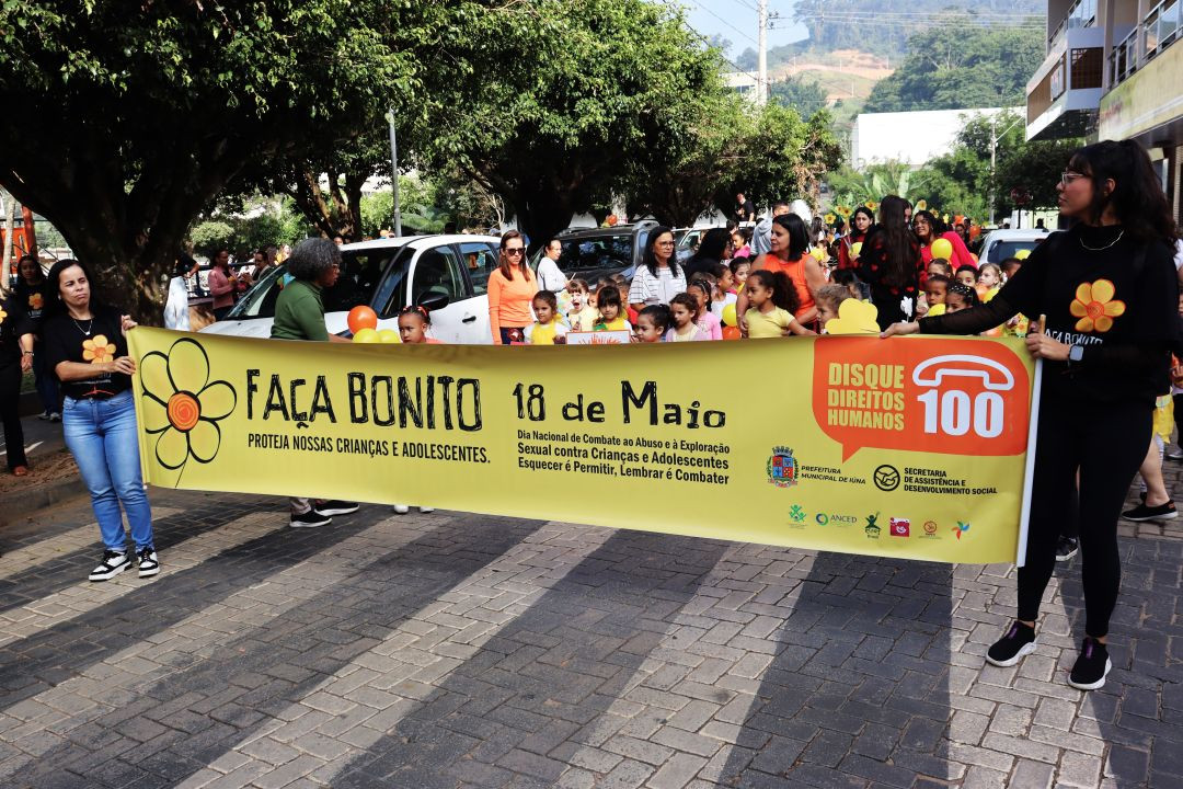 Fotografia colorida horizontal em plano médio mostrando duas mulheres segurando as extremidades de uma grande faixa amarela em uma faixa de pedestres no centro de Iúna. Na faixa, está escrito em letras pretas 