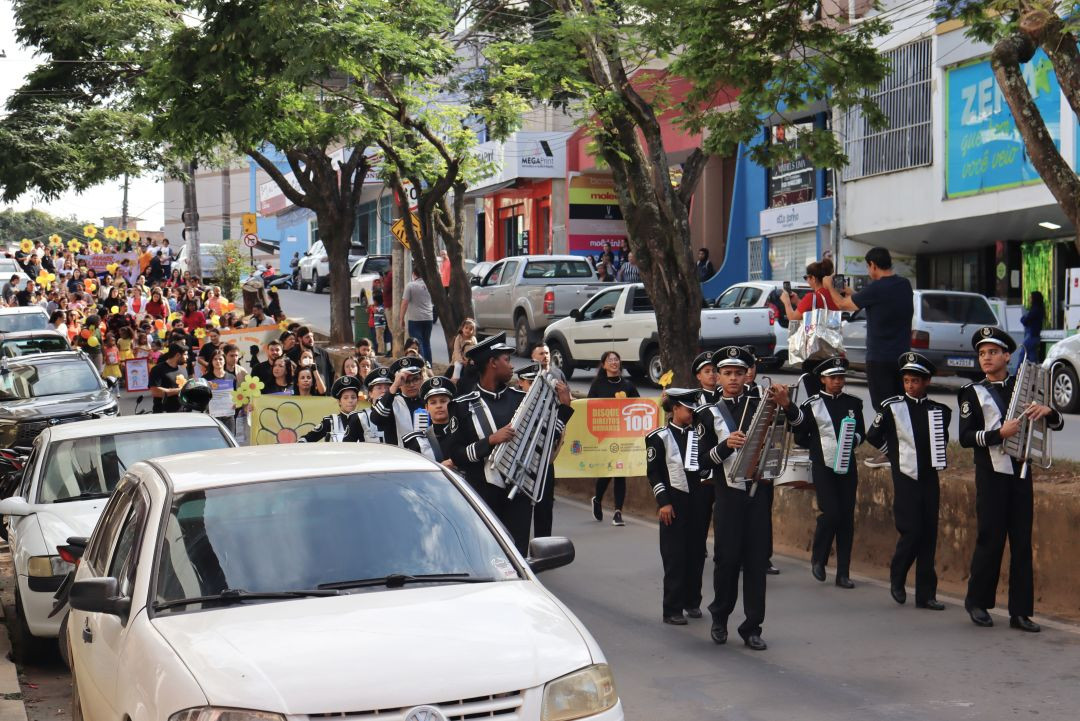 Fotografia colorida horizontal capturada em ângulo paralelo à rua. Em primeiro plano, integrantes da banda marcial juvenil COMUTI marcham vestindo fardamento preto e detalhes prateados, tocando liras e escaletas. Atrás deles, o público da caminhada avança carregando balões e flores amarelas artificiais. Carros estão estacionados ao longo do meio-fio e estabelecimentos comerciais locais compõem o cenário ao fundo.