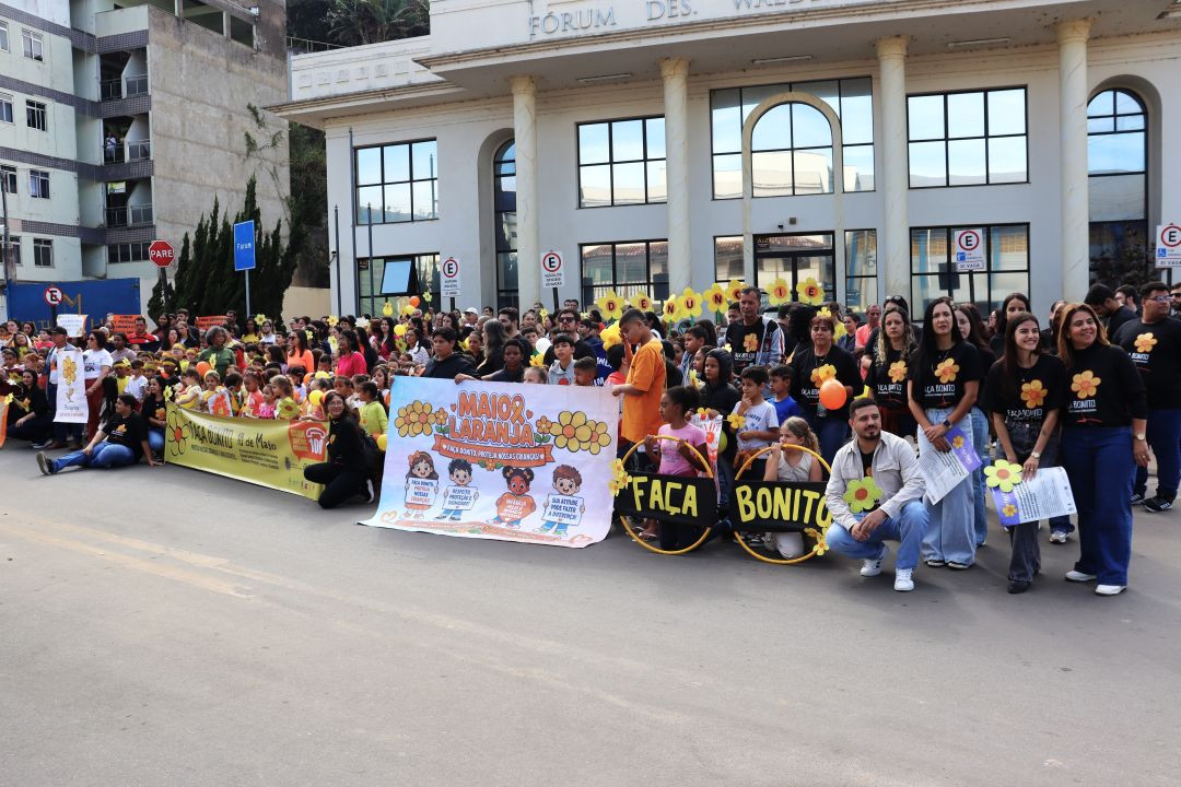 Fotografia colorida horizontal registrando a concentração dos participantes da caminhada reunidos em frente ao prédio do Fórum Desembargador Waldemar Pereira, que possui colunas brancas na fachada. No centro do grupo, adultos e crianças seguram um grande banner com desenhos infantis e o texto 