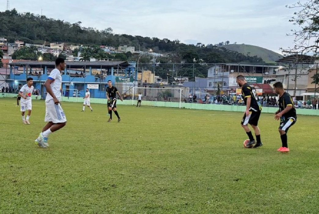 Fotografia em plano aberto de uma partida de futebol de campo durante o dia. No gramado verde, dois jogadores vestindo uniforme preto com detalhes em amarelo dominam e protegem a bola no lado direito. À esquerda, dois jogadores com uniforme branco observam e se posicionam para a marcação. Ao fundo, vê-se a estrutura do campo com alambrados, faixas de patrocinadores do Sicoob, arquibancadas cobertas e uma colina com casas e vegetação sob um céu nublado.