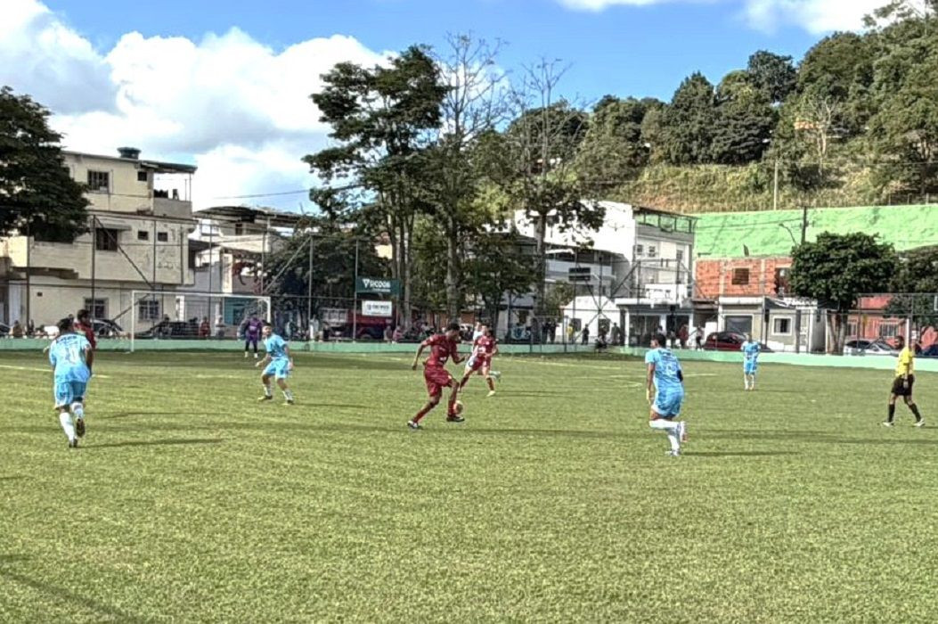 Fotografia de um jogo de futebol em um dia ensolarado. Um jogador com uniforme de cor vinho avança com a bola dominada no centro do gramado, cercado por jogadores com uniforme azul claro espalhados pelo campo. No canto direito, o árbitro da partida acompanha o lance vestindo camisa amarela e calção preto. Ao fundo, destacam-se árvores altas, casas de dois andares e um barranco com vegetação sob um céu azul com nuvens brancas.