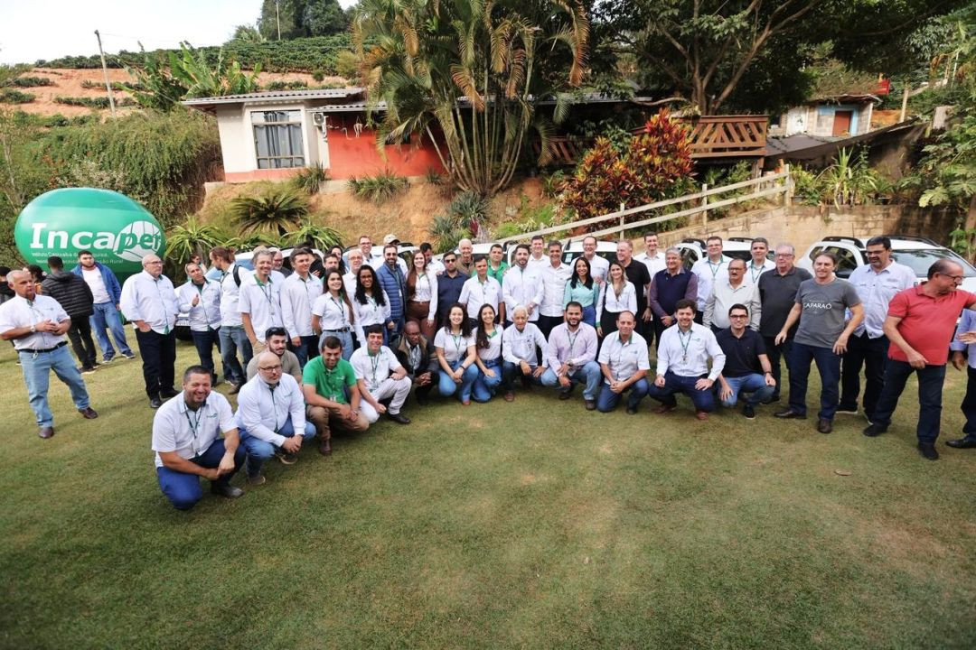 Foto aérea em ângulo aberto mostrando dezenas de participantes do evento reunidos em um gramado em frente a uma casa de campo com telhado colonial. Ao fundo, há vários veículos utilitários brancos alinhados e um balão inflável verde com a marca do Incaper. O grupo é composto por produtores, técnicos e autoridades posando juntos para a fotografia oficial.