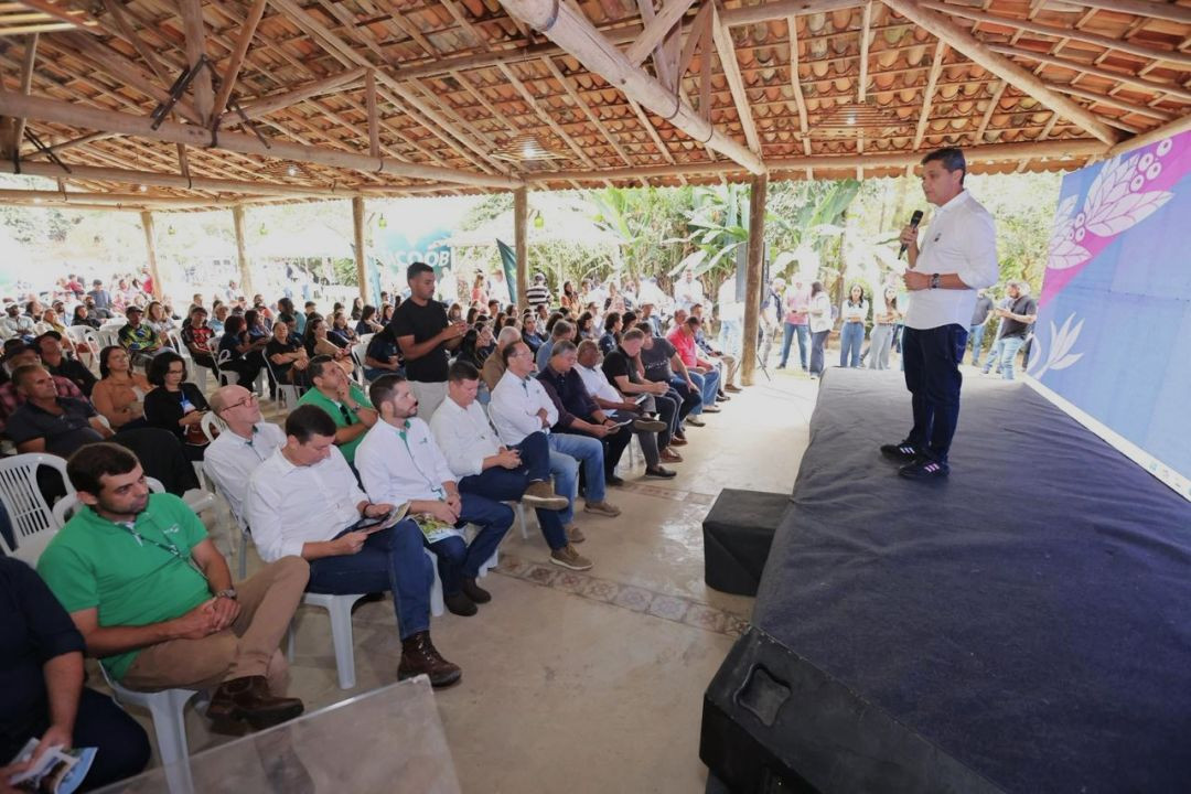 Visão geral do público assistindo à cerimônia dentro de um pavilhão coberto e aberto nas laterais, cercado por vegetação. À direita, sobre um palco baixo, o governador Ricardo Ferraço fala ao microfone voltado para as fileiras de cadeiras ocupadas por produtores, técnicos do Incaper em camisas brancas e verdes, e demais autoridades locais.