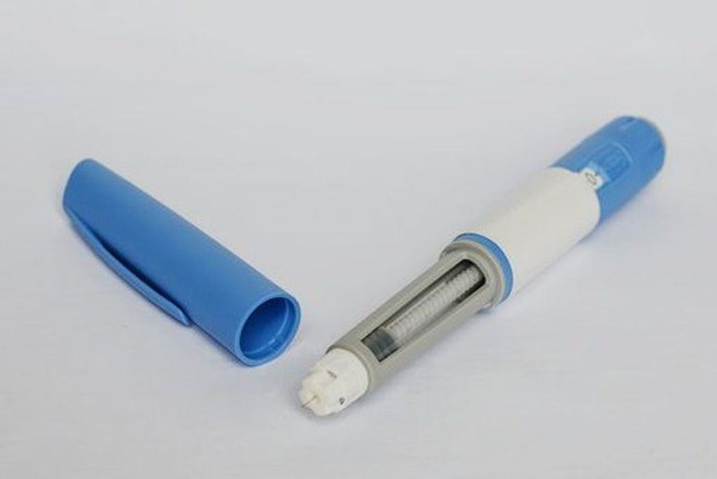 Fotografia em plano detalhe e fundo branco estúdio apresentando uma caneta de aplicação de medicamento injetável desmontada. Do lado esquerdo, vê-se a tampa protetora de plástico azul claro na horizontal. Do lado direito, o corpo principal do aplicador em tons de azul e branco, exibindo o compartimento transparente onde se localiza o êmbolo e o mecanismo de rosca com uma pequena agulha acoplada na extremidade.