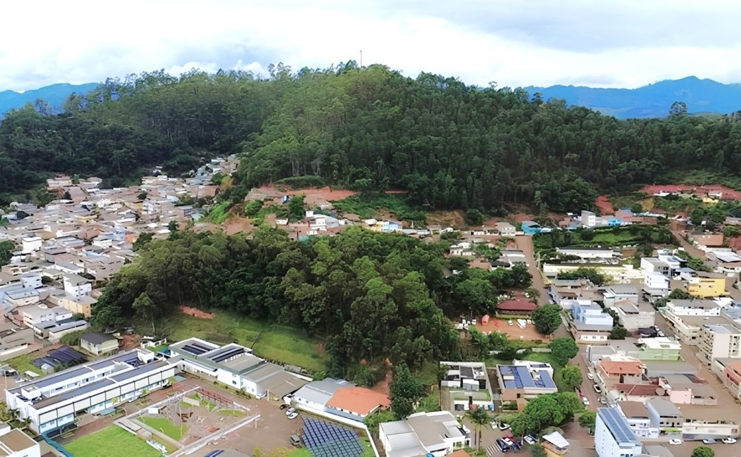 Vista aérea panorâmica de um trecho urbano do município de Ibatiba. No centro e ao fundo, destaca-se uma extensa e densa colina coberta por vegetação nativa de Mata Atlântica, correspondente ao Horto Florestal. Na parte inferior e nas laterais da imagem, observam-se residências, ruas asfaltadas, edificações comerciais e uma escola com quadra externa e placas de energia solar no telhado, mostrando o contraste e a proximidade direta entre a malha urbana e a reserva florestal preservada sob um céu parcialmente nublado.