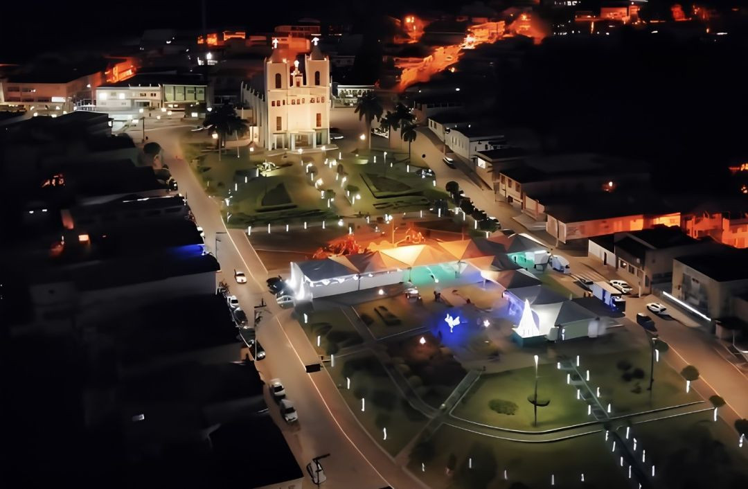 Vista aérea noturna da praça central de São José do Calçado iluminada. Ao fundo, destaca-se a arquitetura da igreja matriz com duas torres brancas iluminadas por refletores. No centro da praça, há uma estrutura de tendas brancas montadas para o evento, com iluminação decorativa em tons de verde, azul e laranja, cercada por ruas adjacentes com alguns carros estacionados.