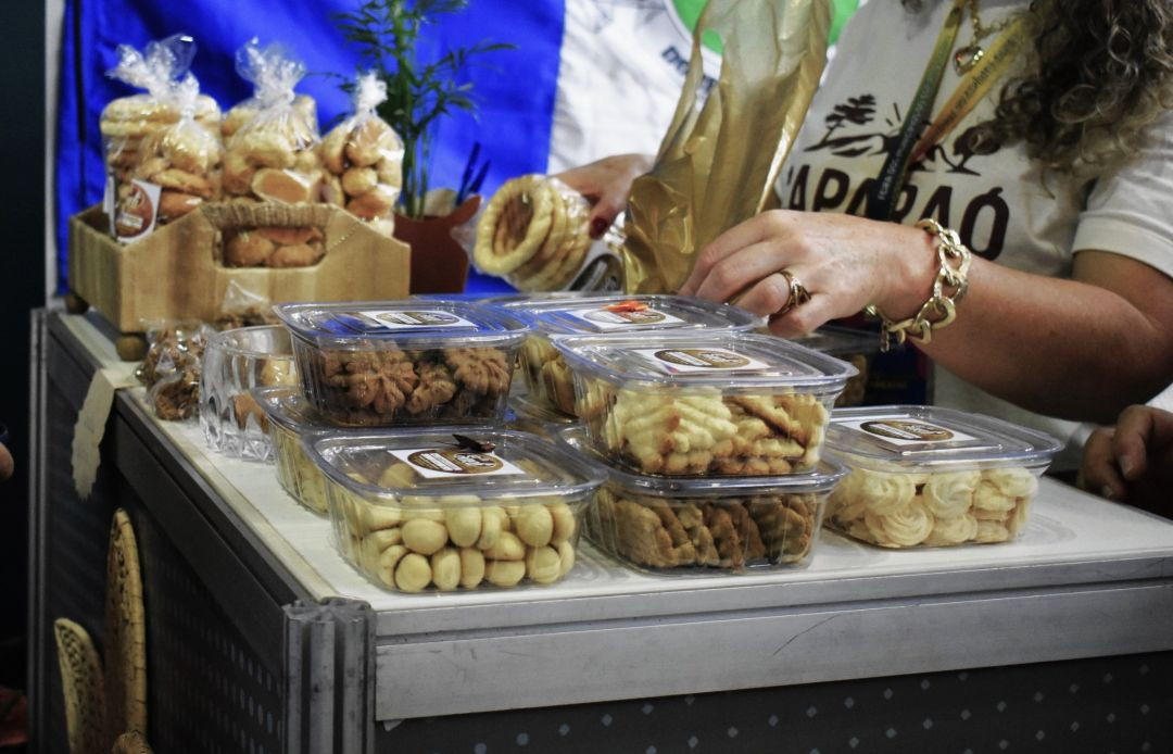 Fotografia em plano detalhe mostrando a exposição de produtos artesanais em um estande. Sobre um balcão cinza, estão organizadas diversas embalagens plásticas transparentes contendo biscoitos e sequilhos de diferentes formatos e tipos. Ao fundo, à esquerda, há pacotes maiores de biscoitos embalados em saquinhos plásticos amarrados com fitas dentro de uma cesta de madeira. À direita, uma mulher usando uma camiseta com a estampa 