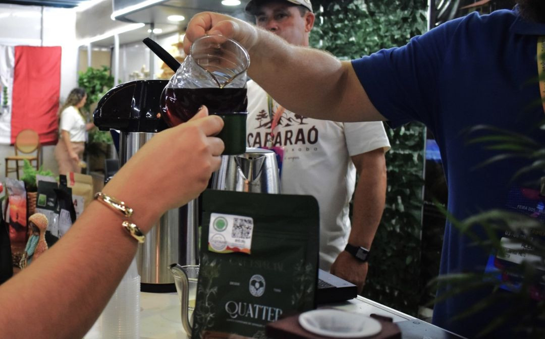Fotografia em plano médio focada na demonstração de café em um estande da feira. À direita, um homem veste uma camiseta branca com a palavra 