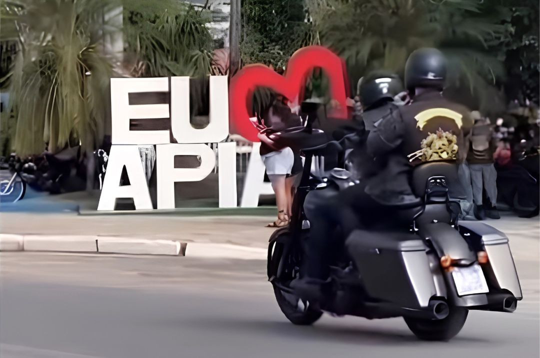 Fotografia em plano médio e de perfil traseiro mostra dois motociclistas, vestindo jaquetas de couro pretas e capacetes escuros, trafegando em uma motocicleta preta de estilo estradeiro (custom/touring) por uma rua asfaltada em Apiacá. Ao fundo, no canteiro central arborizado, destaca-se um letreiro turístico com letras brancas grandes onde se lê 