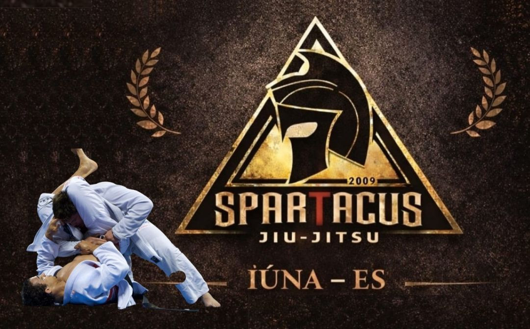 Imagem de dois lutadores com quimonos brancos estão lutando em técnica de chão de Jiu-Jitus e à direita e acima, o logotipo oficial do evento Spartacus Jiu-Jitsu Iúna. A imagem apresenta um fundo texturizado em tons escuros e bronze. No centro, há um grande triângulo dourado com bordas duplas, contendo a ilustração estilizada de um capacete de guerreiro espartano de perfil, voltado para a esquerda. Abaixo do capacete, consta o ano 