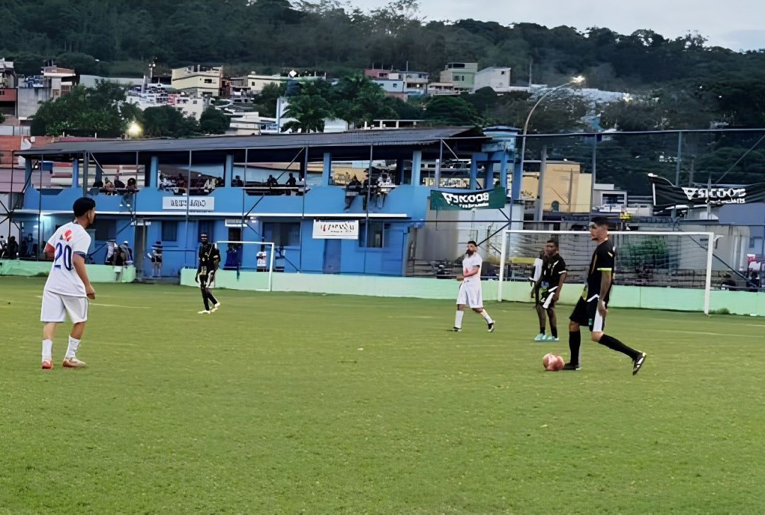 Fotografia em plano aberto de uma partida de futebol de campo durante o dia. No gramado verde, jogadores com uniformes distintos disputam a bola: um jogador à direita, vestindo uniforme preto com detalhes dourados, conduz a bola com o pé direito, enquanto um adversário com uniforme branco (camisa com o número 20 em azul nas costas) o observa à esquerda. Ao fundo, destaca-se uma arquibancada coberta de dois andares pintada de azul claro, com faixas de patrocinadores como o Sicoob. Atrás do estádio, há uma colina com vegetação verde e casas escalonadas típicas de uma região montanhosa.