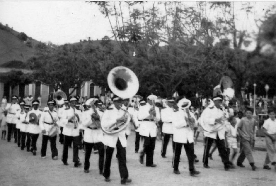 Fotografia antiga em preto e branco mostrando os músicos da Lyra Carlos Gomes desfilando em uma rua de terra batida na década de 1960. Os integrantes vestem uniformes brancos de gala com quepes e calças escuras, tocando instrumentos de sopro como sousafones, trompetes e saxofones. Ao fundo, árvores e o relevo montanhoso da região.