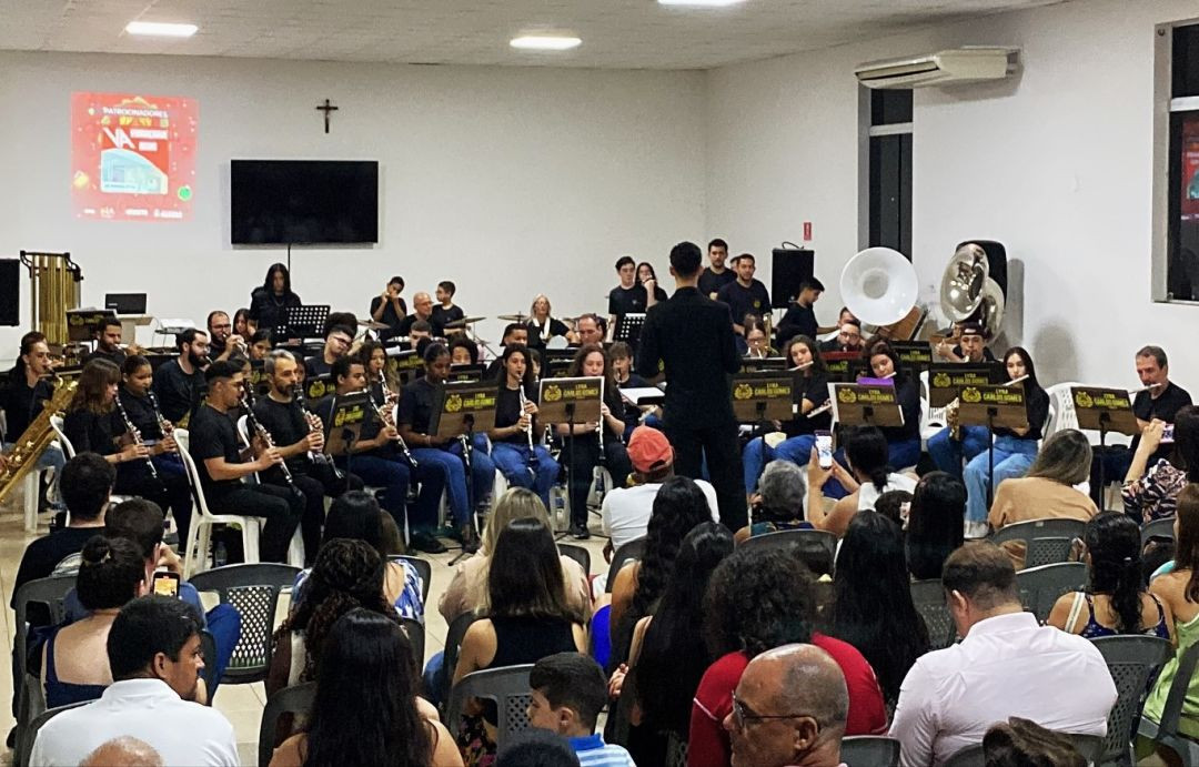 Foto colorida atual de uma apresentação da Lyra Carlos Gomes em um auditório fechado e iluminado. Os músicos estão sentados em semicírculo, vestindo camisas pretas e calças jeans, tocando instrumentos de sopro e percussão. Ao centro, de costas, o maestro rege o grupo diante de uma plateia sentada que assiste ao concerto.