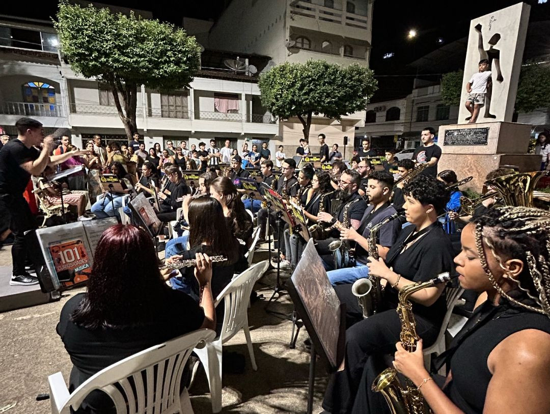 Fotografia colorida tirada à noite em uma praça de Alegre, mostrando a banda disposta em cadeiras realizando uma apresentação pública. Em primeiro plano, detalhes do naipe de saxofones e flautas transversais. Ao fundo, o público assiste em pé ao redor do monumento da praça iluminada.