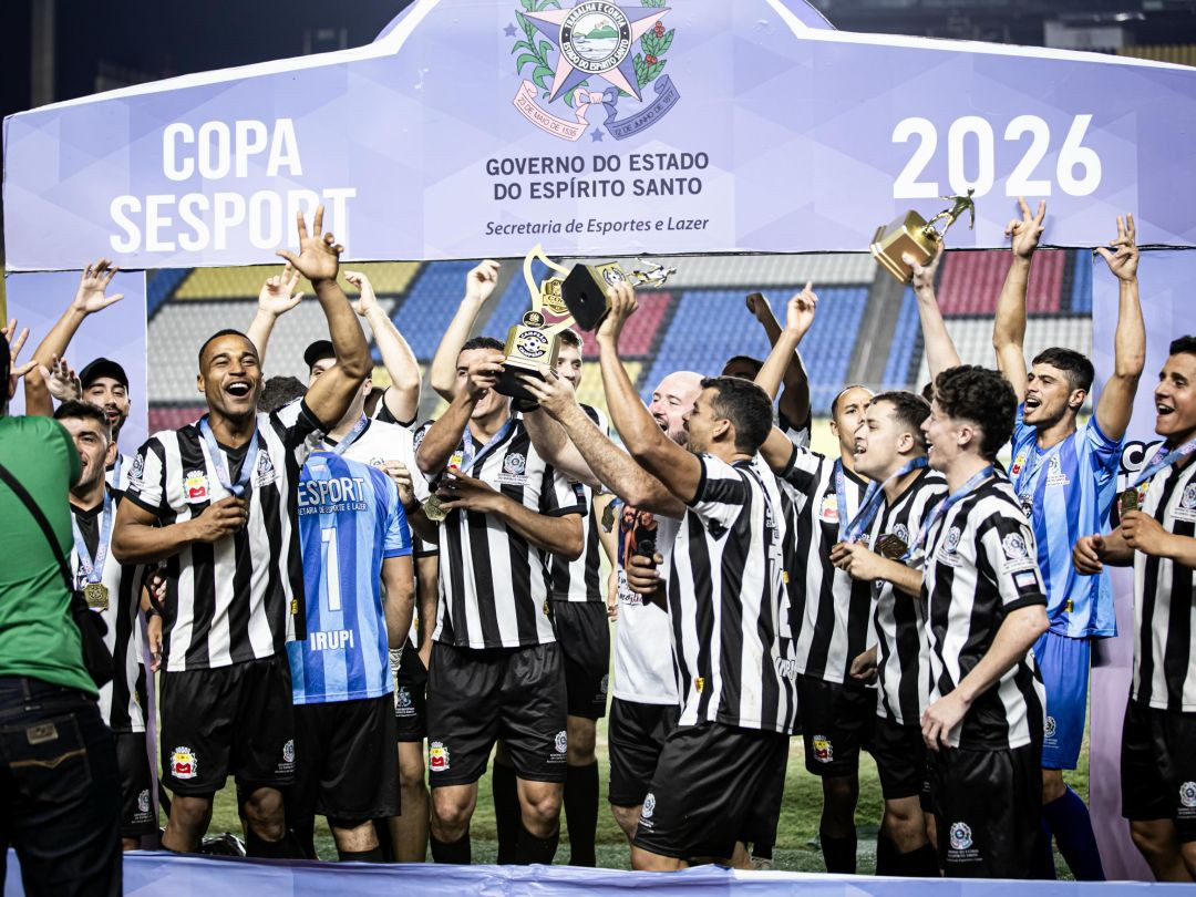 Fotografia horizontal do momento de celebração da equipe de Irupi sob o portal oficial da Copa Sesport do Governo do Estado do Espírito Santo. Atletas com uniformes listrados em preto e branco e a comissão técnica aparecem com os braços levantados, sorrindo e gritando em comemoração, enquanto erguem a taça de campeão e os troféus individuais no gramado.