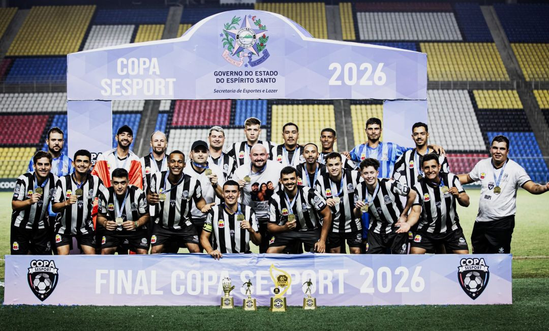 Fotografia oficial da equipe de Irupi posando sob o pórtico de campeão no gramado do Estádio Kleber Andrade. Ao fundo, uma estrutura cenográfica azul traz o brasão do Governo do Estado do Espírito Santo, a inscrição 