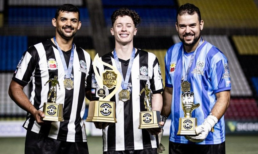 ): Fotografia em plano médio de três integrantes da equipe de Irupi no gramado do Estádio Kleber Andrade à noite. À esquerda, um jogador com uniforme listrado em preto e branco e medalha no pescoço segura o troféu de artilheiro. Ao centro, outro jogador com o mesmo uniforme sorri segurando a taça dourada de campeão da Copa Sesport. À direita, o goleiro posa com uniforme azul listrado, luvas brancas e o troféu em formato de luva dourada de melhor goleiro.
