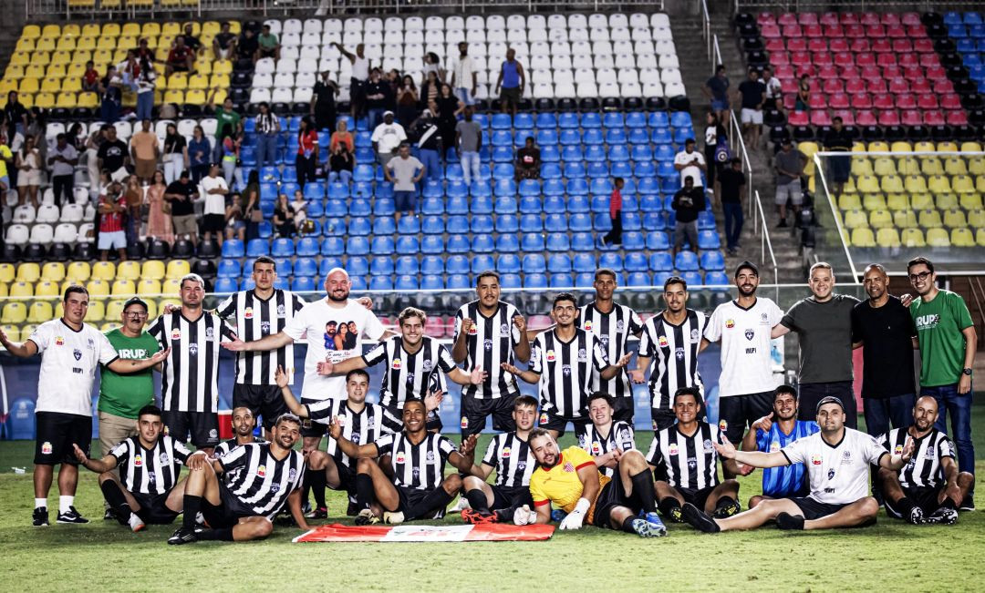 Fotografia panorâmica do elenco completo e da comissão técnica de Irupi reunidos no gramado, posicionados em frente à arquibancada do estádio, onde torcedores observam sentados nas cadeiras azuis, brancas e amarelas. A equipe está dividida entre atletas de pé e sentados diretamente na grama. No centro, no chão, uma bandeira vermelha e branca está estendida. Os integrantes vestem os uniformes oficiais de jogo, camisas de comissão técnica e camisetas brancas com a estampa do município de Irupi, todos sorrindo de braços abertos em clima de celebração pelo título.
