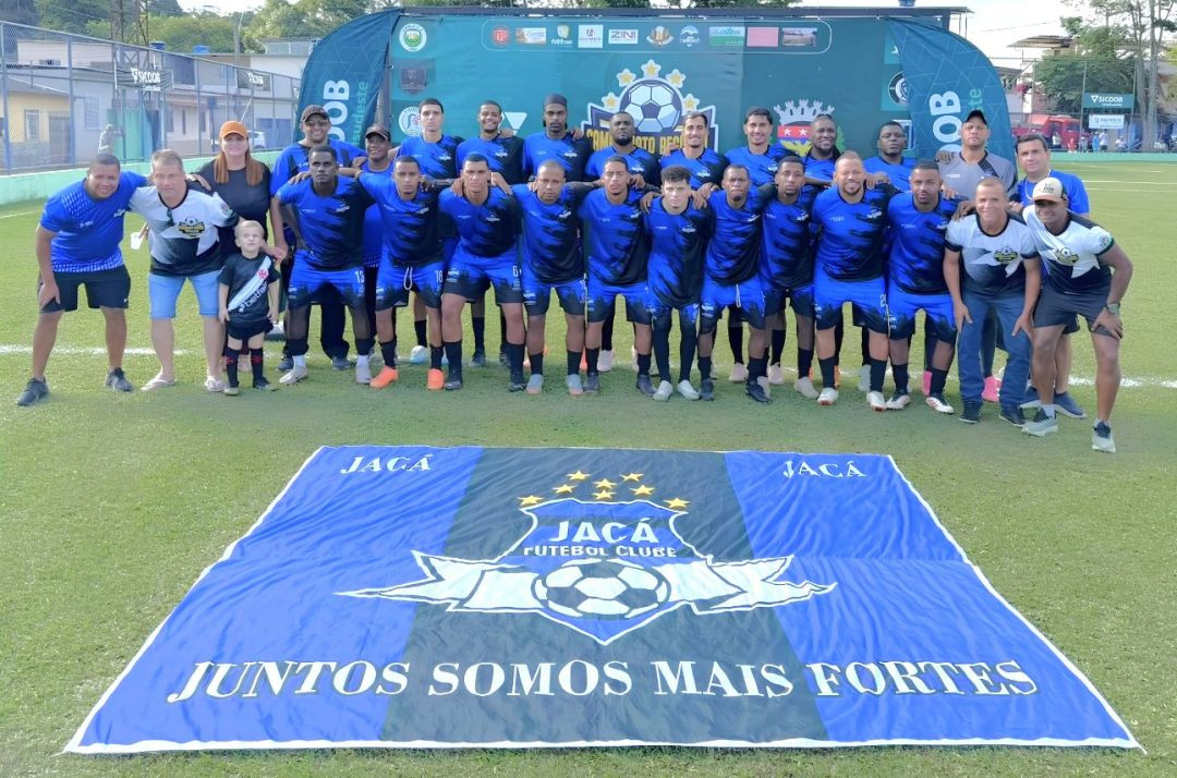 Foto oficial da equipe do Jacá Futebol Clube. Cerca de vinte e cinco atletas e comissão técnica posam perfilados no gramado, vestindo uniforme azul e preto. À frente deles, estendida no chão, está uma grande bandeira azul com o escudo do clube no centro e a frase 