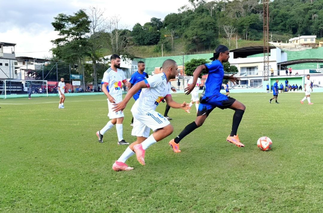 Imagem de jogo em andamento. Um jogador com uniforme azul corre com a bola sob o domínio do pé direito, sendo marcado de perto por um atleta de uniforme branco. Ao fundo, outros jogadores observam o lance no gramado verde sob céu nublado.