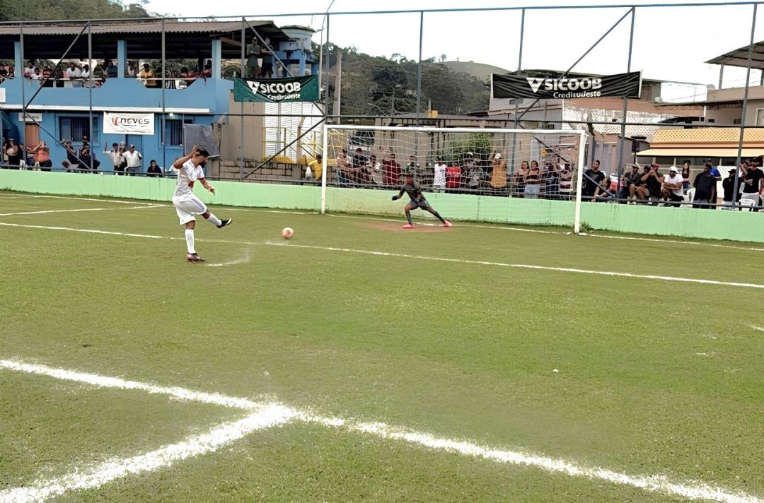 Momento de uma cobrança de pênalti. O cobrador, com uniforme branco, aparece de costas chutando a bola em direção ao gol. O goleiro, de uniforme escuro, está posicionado no centro da meta. Torcedores assistem à cobrança atrás da grade de proteção do estádio.