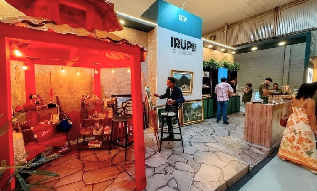 Fotografia colorida do estande do município de Irupi na Feira dos Municípios. À esquerda, destaca-se uma estrutura cenográfica de madeira pintada de vermelho vivo, simulando o quiosque com telhado rústico, iluminada por lâmpadas quentes penduradas. No interior dessa estrutura, há prateleiras e balcões exibindo produtos artesanais, bolsas e pequenos objetos. Ao centro, um homem de paletó escuro está sentado em uma banqueta alta preta, de perfil, pintando em uma tela apoiada em um cavalete de madeira. Atrás dele, há uma parede branca com a inscrição 
