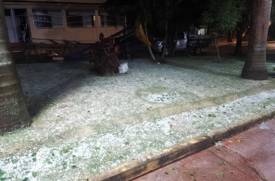 Fotografia noturna de um espaço público gramado no distrito de Itaici, em Muniz Freire. O gramado entre duas palmeiras e próximo ao meio-fio está totalmente coberto por uma camada esbranquiçada de pedras de granizo. Ao fundo, vê-se a fachada de um imóvel residencial com grades e um veículo utilitário estacionado.