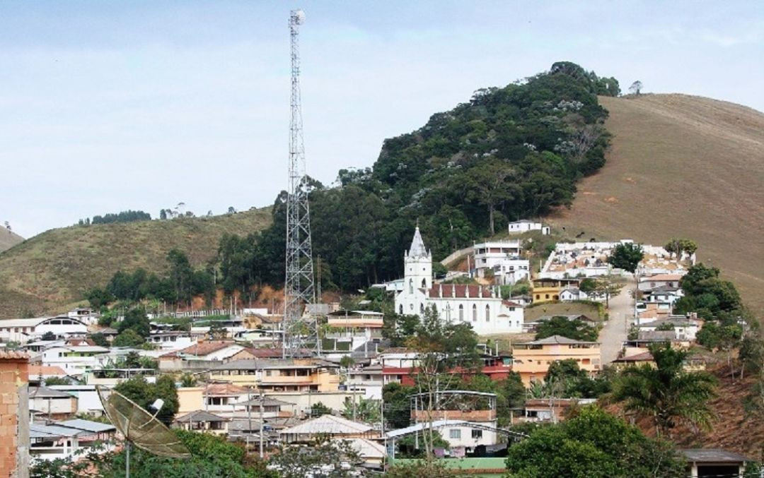 Imagem em plano médio da Paróquia Divino - Sagrado Coração de Jesus. A igreja possui fachada pintada em tom verde-claro com detalhes brancos, janelas em formato ogival (estilo gótico) e uma torre centralizada com uma cruz no topo. À esquerda e ao fundo, observam-se construções residenciais, muros e a encosta de um morro coberto por vegetação rasteira sob céu com nuvens.