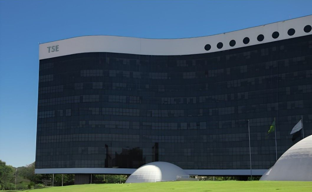 Fachada do edifício do Tribunal Superior Eleitoral (TSE) em Brasília, exibindo arquitetura moderna com linhas onduladas, vidros escuros reflexivos e a sigla TSE fixada no topo esquerdo, sob céu azul limpo.