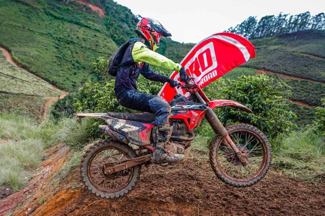 Fotografia em plano médio e diagonal de um motociclista em movimento durante uma trilha de terra em Muniz Freire. O piloto veste equipamentos de proteção nas cores preta e verde-limão, capacete colorido e bota com marcas de lama. A motocicleta, modelo de trilha na cor vermelha, está ligeiramente inclinada sobre um barranco de terra escura e úmida. Ao fundo, uma encosta com plantações verdes e estradas rurais de terra faz o contorno da paisagem montanhosa. Uma bandeira promocional vermelha com escrita branca aparece parcialmente atrás da moto.