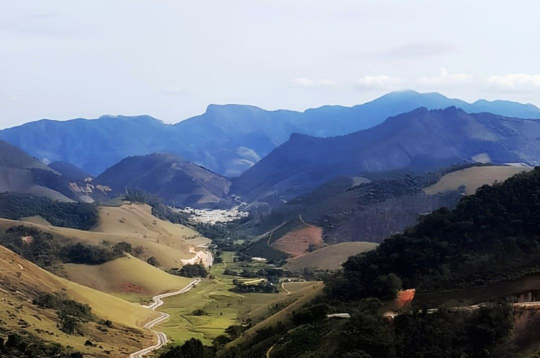 A imagem captura uma vasta paisagem de montanhas onduladas em camadas, com a cadeia montanhosa mais alta ao fundo exibindo uma tonalidade azulada profunda e suavemente envolta em névoa atmosférica. No plano médio, uma vila compacta de telhados brancos se aninha no vale profundo. Uma estrada estreita e sinuosa de asfalto branco serpenteia pelas colinas gramadas, de cor verde-oliva e bronzeada, em direção à vila. O primeiro plano apresenta colinas mais próximas com vegetação densa e escura. O céu está claro e parcialmente nublado.