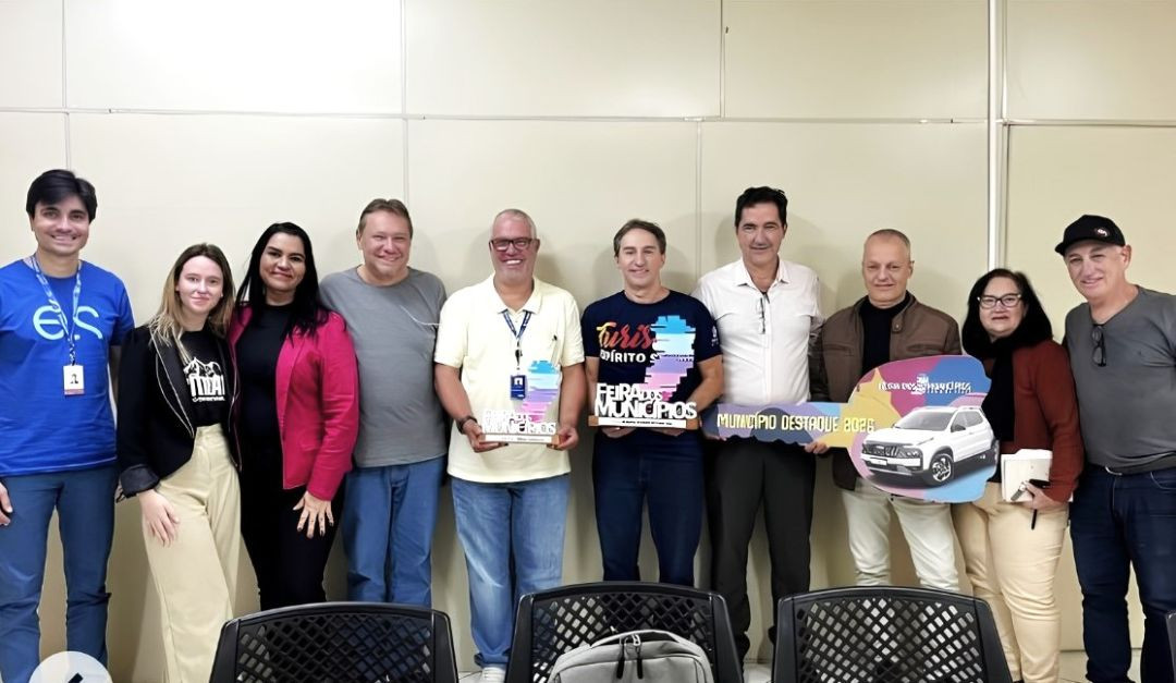 Grupo de dez pessoas, homens e mulheres, posados lado a lado em pé em uma sala de reuniões com parede clara ao fundo. No centro, três homens seguram premiações da Feira dos Municípios, incluindo um troféu em formato do mapa do Espírito Santo e uma chave cenográfica grande com a frase 