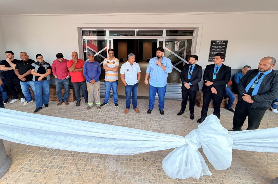 Homens e mulheres perfilados em frente à entrada da Capela Mortuária de Iúna durante a solenidade. No centro, o prefeito Romário fala ao microfone. Em primeiro plano, uma grande fita de tecido branco esticada para o corte inaugural.