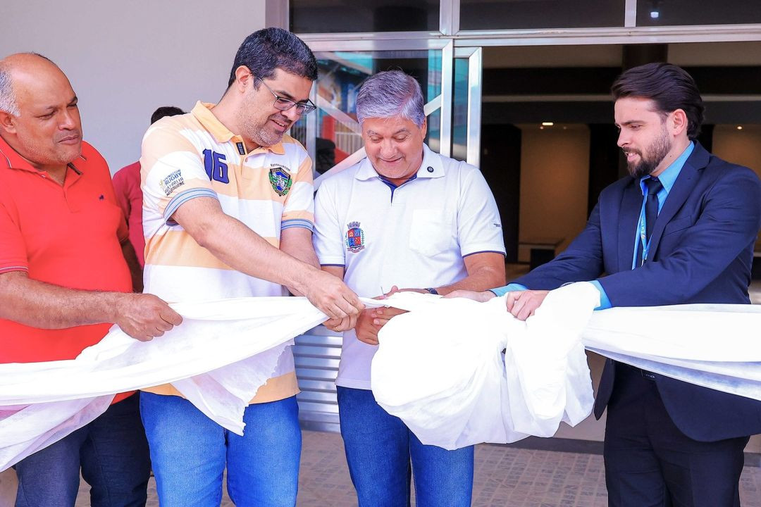Quatro homens segurando e cortando a fita branca inaugural do espaço. Da esquerda para a direita, um homem de camisa vermelha, um de camisa listrada amarela e branca, um de camisa polo branca com o brasão do município e um de terno escuro.