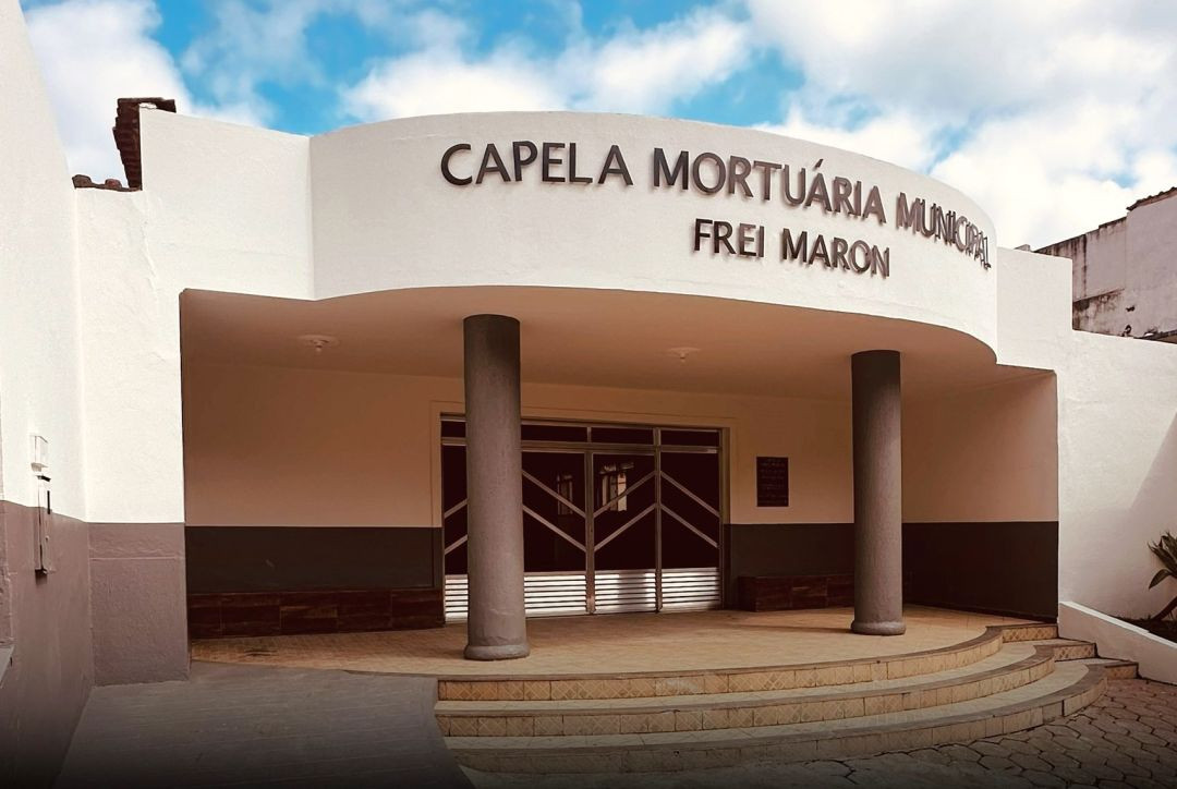 Fachada externa da Capela Mortuária Municipal Frei Maron em Iúna. O prédio tem paredes brancas, detalhes em cinza escuro, marquise arredondada sustentada por duas colunas cilíndricas cinzas e portas de correr de metal. O céu ao fundo está parcialmente nublado.