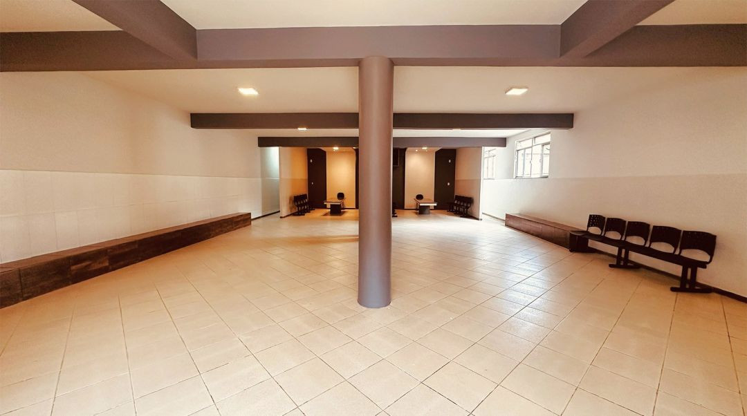 Vista do interior amplo da capela revitalizada. O piso é revestido com pisos cerâmicos claros e há uma coluna cinza central que sustenta as vigas do teto. Ao fundo, veem-se as entradas para as duas salas de velório e, nas laterais, longos bancos de madeira e cadeiras pretas fixadas na parede.