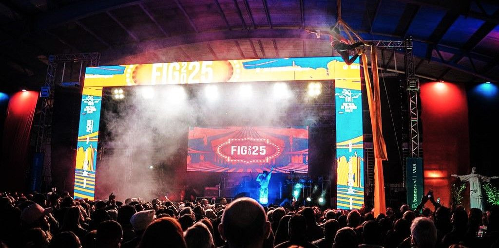 Fotografia horizontal de um palco sob uma estrutura de galpão coberto. O portal do palco exibe uma identidade visual colorida em tons de azul, amarelo e laranja, com a inscrição 
