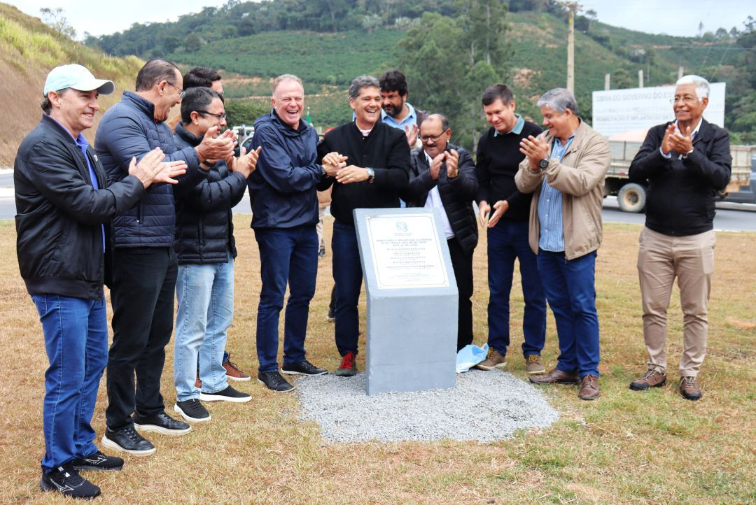 Grupo de homens em pé em uma área gramada ao ar livre, aplaudindo ao redor de um marco de concreto cinza com uma placa de metal branca instalada. Entre eles, o governador Ricardo Ferraço, de jaqueta escura, sorri e cumprimenta os presentes. Ao fundo, uma encosta verde com plantações de café e um caminhão na rodovia asfaltada.