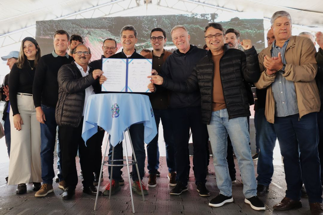 Registro oficial no palco principal sob uma tenda. O governador Ricardo Ferraço, o ex-governador Renato Casagrande e prefeitos da região seguram juntos um documento oficial aberto dentro de uma pasta azul sobre uma mesa redonda coberta com uma toalha azul clara com o brasão do estado. Todos sorriem para a foto.