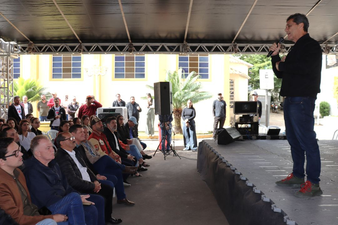 Visão lateral do palco de eventos montado em praça pública. O governador Ricardo Ferraço está em pé no palco à direita, segurando um microfone e discursando para o público. À esquerda, moradores e autoridades locais assistem sentados em cadeiras de plástico sob a cobertura da tenda.
