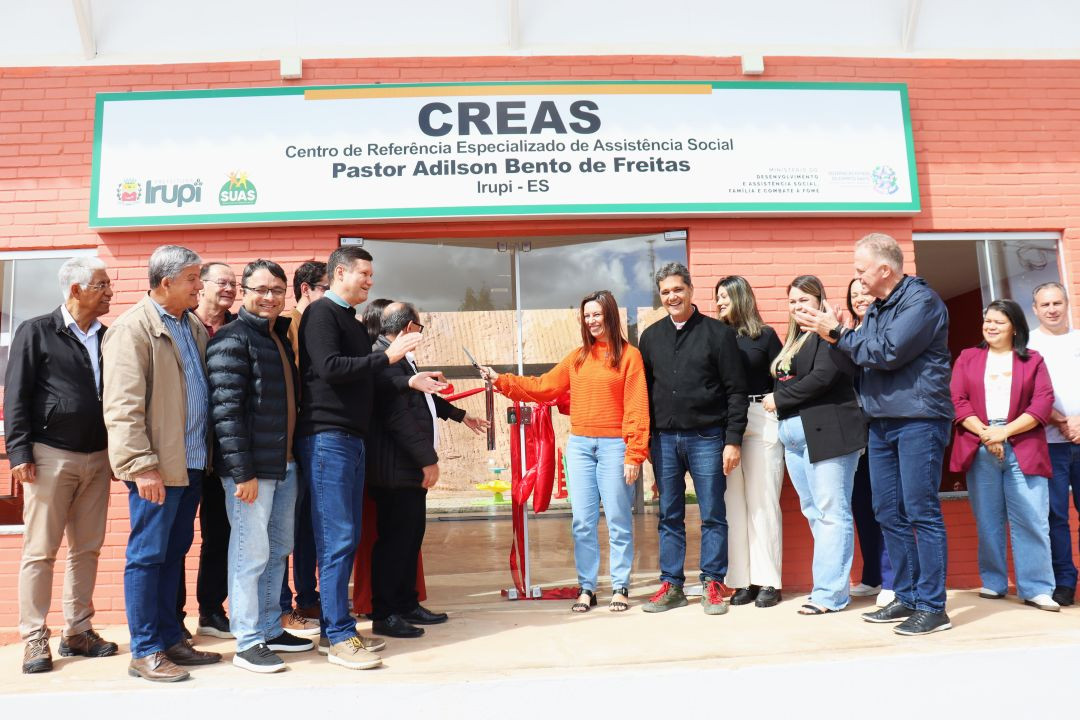 Momento do corte da fita inaugural vermelha na entrada do novo prédio do CREAS de Irupi. Uma mulher de blusa laranja segura a tesoura ao centro, cercada por homens e mulheres que aplaudem a ação. Acima da porta de vidro, há uma placa verde e branca com a inscrição 