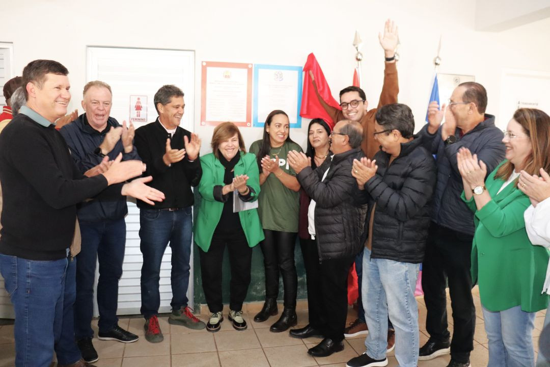 Autoridades e lideranças comunitárias reunidas em um ambiente interno de piso claro, sorrindo e batendo palmas em frente a duas placas inaugurais fixadas na parede branca ao lado de uma porta. Duas mulheres vestem blazers na cor verde e homens usam jaquetas de inverno.