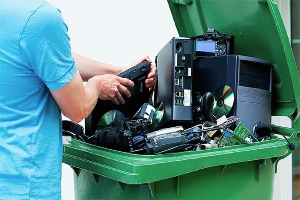 Homem de perfil, vestindo camisa azul, coloca um notebook antigo dentro de uma lixeira grande de plástico verde, que está aberta e cheia de lixo eletrônico. No interior do contêiner, são visíveis mídias de CD e DVD, gabinetes de computador, placas de circuito impresso, câmeras fotográficas e fones de ouvido acumulados para descarte.