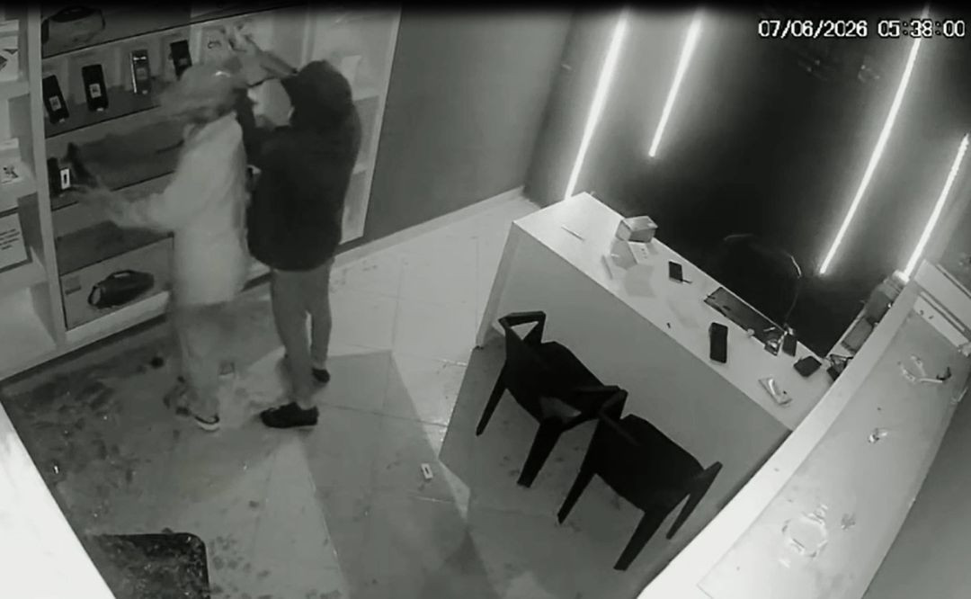 Imagem em preto e branco de câmera de segurança interna do estabelecimento, datada de 7 de junho de 2026, às 05:38. Dois homens encapuzados e com bonés aparecem recolhendo aparelhos celulares de prateleiras no interior da loja. O chão está coberto por estilhaços de vidro e restos da porta arrombada. À direita, há um balcão de atendimento branco com duas cadeiras escuras.