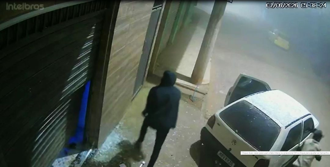 Imagem de câmera de segurança noturna na madrugada de 7 de junho de 2026, às 05:38. Um homem de costas, vestindo casaco escuro com capuz, caminha em direção à porta de aço retorcida e arrombada de um comércio. Ao lado direito, um veículo Volkswagen Gol branco está estacionado na rua com a porta do motorista aberta. Outro homem aparece parcialmente no canto inferior direito.