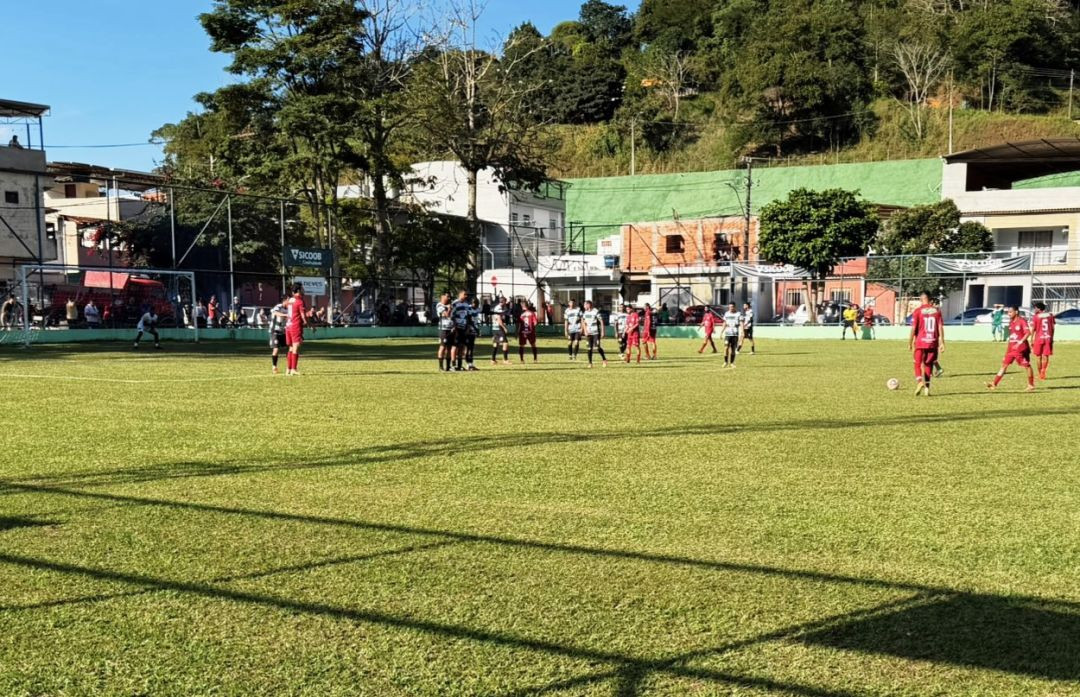 Vista panorâmica do gramado do Estádio Municipal de Dores do Rio Preto em um dia ensolarado. Jogadores das equipes de uniforme listrado em verde e branco e uniforme grená estão posicionados no campo durante uma cobrança de falta ou paralisação. Ao fundo, veem-se alambrados, faixas de patrocinadores como o Sicoob, árvores e construções da cidade próximas a um morro.