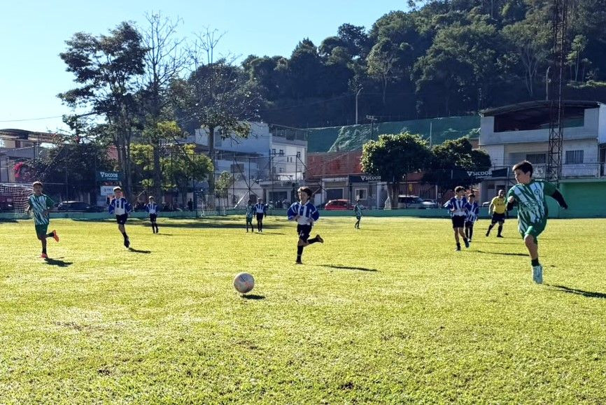 Fotografia de um jogo de futebol infantil em andamento durante um dia ensolarado. Vários meninos correm pelo gramado verde atrás da bola oficial que está no centro do campo. Uma equipe veste uniforme listrado em azul e branco, e a outra veste uniforme listrado em verde e branco. Ao fundo, vê-se árvores, casas da cidade e placas de patrocínio do Sicoob.