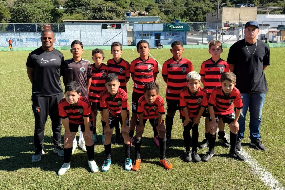 Foto oficial posada da equipe Sub-11 de Dores do Rio Preto no campo de futebol. Os dez jogadores mirins vestem uniformes com listras horizontais vermelhas e pretas. Dois técnicos adultos ladeiam as crianças, um à esquerda vestindo camisa preta da Nike e outro à direita com camisa preta lisa e boné. Ao fundo, o gramado e o muro do estádio sob a luz do sol.
